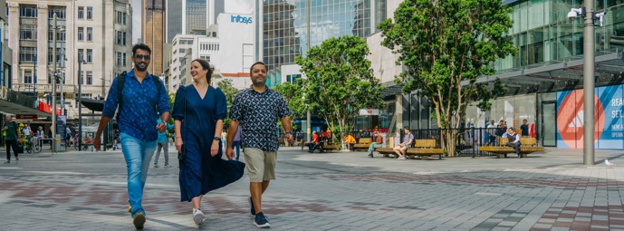 Friends walking on Te Komititanga square and behind them is the city centre and Queen St